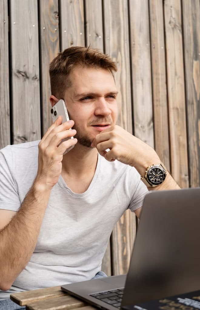 Homme au téléphone devant son ordinateur portable, adossé à une paroi de bois